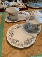 Three sets of fine china teacups with matching snack plates, detailed floral patterns. First set has black and gray leaf vine design.
