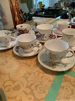 Photo showing assorted fine china teacups and saucers arranged on a tablecloth with a gold pattern, highlighting varied floral patterns and gold trims.