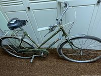 Side and angled views of a vintage Raleigh step-through bicycle in olive green with white grips, black saddle, rear rack, chain guard, and fenders.