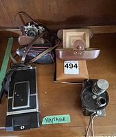 Vintage cameras and leather cases on wooden surface including Revere Ventura camera and vintage handheld movie camera.