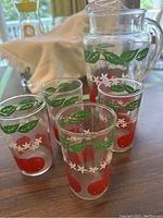 Photo showing four small glasses and the pitcher with red tomato, green leaves, and white floral designs.