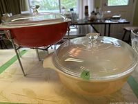 Photo showing both casseroles together: Pyrex dish in dusty rose on heat frame at back and Fire King casserole in pearlized orange in front.