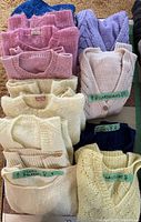 Top view showing ten folded hand knit sweaters in pastel colors including pink, blue, purple, and yellow. Some sweaters are labeled with tags indicating types such as 'cardigans' and 'long sleeves'.