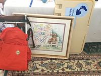 The full lot setup on patterned rug showing the red REI hiking backpack, framed Helen Paul print resting against the wall, and beige Koala baby changing station behind.