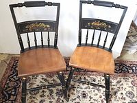 Two Bent Bros black lacquered dining chairs with natural wood seats and floral stencil decoration on backs, placed on patterned carpet.