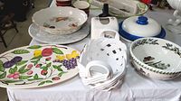 Overview of dinnerware, bakeware, kitchenware and serveware on white tablecloth including bowls, casserole dishes, tissue box cover, baking dishes, tea kettle and woven baskets.