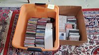 Photo of orange bin filled with stacked music CDs and white Bose Wave Radio on top, next to cardboard box containing cassette tapes and additional CDs.
