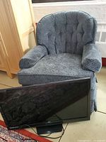 Gray upholstered swivel rocking chair with tufted backrest and cushioned armrests, positioned behind a flat screen TV.