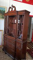 Front view of the tall mid-century china cabinet hutch showing glass-paneled upper doors with decorative wooden framework and carved top crest.