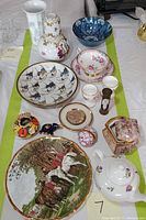 Full view of all porcelain items laid out together on a white cloth showing vases, decorative plate, teacups, saucer, small cups, hourglass timer, and figurines.