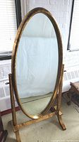 Full view of the oval cheval floor mirror with gold enameled wooden frame and swivel base on the ground.