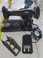 Side angled view of black vintage Singer sewing machine with gold detailing alongside black foot pedal and ornate cast iron napkin holder in front.