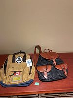 Two backpacks side by side on a wooden surface; tan Red Canoe Heritage Bell Canada lineman bag and black and brown two-pocket leather-trimmed backpack