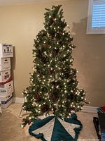 Full view of the assembled 7 ft tall artificial Christmas tree decorated with lit white lights, pinecones, and berries, with tree skirt at the base.
