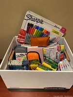 Top view of a white bankers box filled with assorted arts and crafts supplies including Sharpie markers, fabric markers, colored pencils, crayons, and various stationery items.