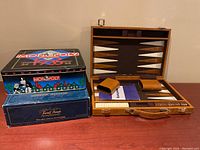 Photo showing backgammon set open with wooden case, felt playing surface, and two leather dice cups, alongside Monopoly 1935 Commemorative Edition tin and Trivial Pursuit box.