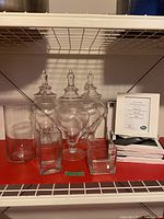 Photo showing three large pointed lid apothecary glass candy jars and multiple smaller glass containers on red shelf along with boxed Umbra white picture frames