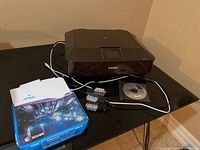 Canon Pixma MG7120 printer with owner's manual booklet, extra ink cartridges in packaging, a CD installation disc, and a bundle of Xerox Versatile multipurpose printer paper on a black table.