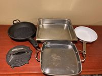 Set of cookware displayed on wooden table including cast iron pans, bacon press, and stainless steel pans.