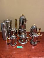 Polished stainless steel coffee set including multiple pots and creamers with gold accents, alongside two stainless steel thermoses, all placed on wooden table surface
