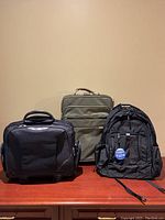 Photo showing front view of three items: olive green suitcase, black briefcase, and black backpack arranged side by side.