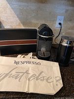 Nespresso Pixie espresso machine next to Aeroccino milk frother, Barista apron box, and cloth tote bag on a kitchen counter.