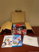 Opened box showing various cables, two Wii remotes, and manual laid out with two Wii game cases in front.
