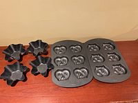 Two Wilton pretzel baking pans each with six pretzel-shaped molds and four black metal fluted baking bowls for shaping wraps/tacos.