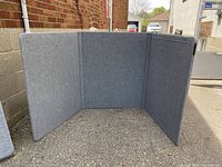 Front view of the tri-fold exhibit display standing open on asphalt against a brick wall, showing grey fabric covering and three connected panels.