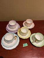 Photo of all 5 pastel colored Royal Albert tea cups with matching saucers on a wooden surface, showing gold trim and scalloped edges.