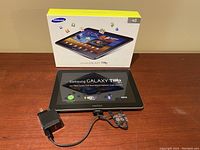 Samsung Galaxy Tab 10.1 tablet with protective film, original box, charger and USB cable on wooden surface.