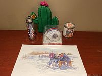 Photo showing all items on wood surface: cactus golf club head cover, water bottle with PGA Tour golf tees, signed mini football helmet, T-shirt in can, and print.