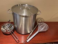 Full view of stainless steel pot with lid and two metal strainer spoons on wooden surface.