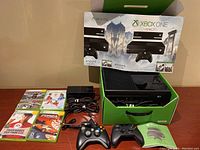Xbox One console in box with Kinect sensor, two black wireless controllers, power supply with cables, and four Xbox 360 game cases arranged on table.
