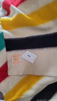 Close-up photo showing the wool blanket's multi-colored stripes, including red, yellow, green, and black on a white background, along with the manufacturer's label confirming 100% wool and made in England.