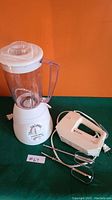 White Proctor Silex blender and white Black & Decker hand mixer with beaters and dough hooks included, shown on green surface with orange background.