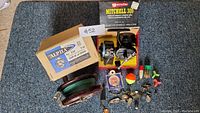 Wide photo of fishing gear showing Mitchell 300 spinning reel in box, Alpha solid cast bracket box, fishing line spool, assorted lures and bobbers, fish hooks scattered on carpet.