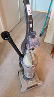 Front and slightly top-angle view showing the full upright vacuum with dust container and attached brush hose accessory.