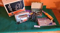 Photo of the Black and Decker corded jigsaw, set of blades, manual stapler with staples in open box, putty knife with orange handle, emergency candles box and user manual on a green felt surface, showing full lot contents.