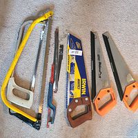Photo showing 6 saws laid out on a carpeted floor, including hacksaws and handsaws.