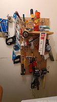 Wooden pegboard with hand tools including screwdrivers, saws, tape, a can of lubricant, small hand rake, and hammer.