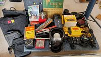 Overview of various vintage photography and darkroom equipment items arranged on table including bags, film rolls, lens components, filters, safelight boxes, paper trays, and cables.