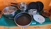 Photo showcasing multiple kitchen cookware items including frying pan, roasters, pots with lids, sieve, and steamer parts all arranged on green cloth.