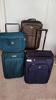 Three pieces of luggage: a green duffel bag stacked on a blue suitcase, and a brown suitcase behind them. All have wheels and pull handles.
