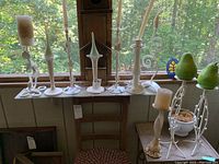 White metal candle holders of various shapes and sizes on a wooden bench near window, with a decorative flower pot containing two artificial pears on a metal stand.