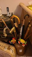 View of multiple decorative and novelty canes, walking sticks, an umbrella cane, and colorful wooden children's craft toys all gathered inside a brown container.