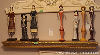 Seven early 20th century styled wooden lady figures, hand-painted, displayed on a decorative shelf around 48 inches wide.