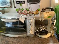 View of all three appliances: Hamilton Beach mixer, Aroma rice cooker in box, Oster toaster oven with beaters and power cords visible
