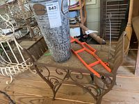 Side view of wrought iron bench seat showing detailed scroll and leaf metalwork and weathered metal plant bucket placed on top.