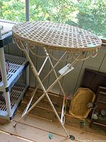 Metal patio table with oval lattice top shown fully assembled and standing on a wooden floor inside a balcony or porch area. Weathered paint finish visible on metal.
